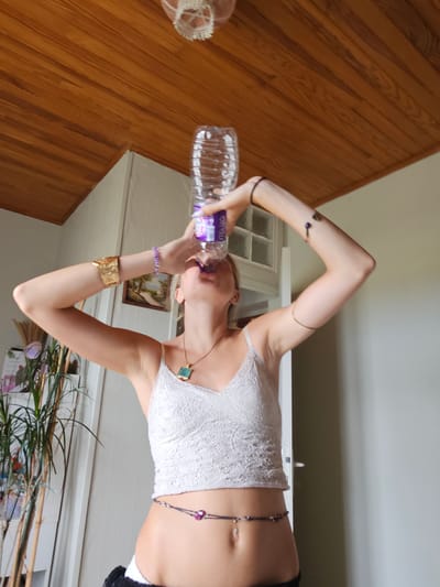 A young woman with blonde hair is captured from the chest up in a low-angle shot, holding a clear plastic water bottle above her head and drinking from it. She is wearing a white lace camisole, a gold cuff bracelet on her left arm, a purple beaded bracelet on her right wrist, a blue pendant necklace, and a purple beaded belly chain. Her arms are bent at the elbows, and her hands are positioned to hold the bottle. The background features a wooden ceiling with horizontal planks, a white wall, and a closed door. To the left, a tall potted plant with green stalks is visible. The overall lighting suggests an indoor setting with natural light coming from an unseen source. The image provides no information about a specific location in Saint-Germain-de-Lusignan, France.