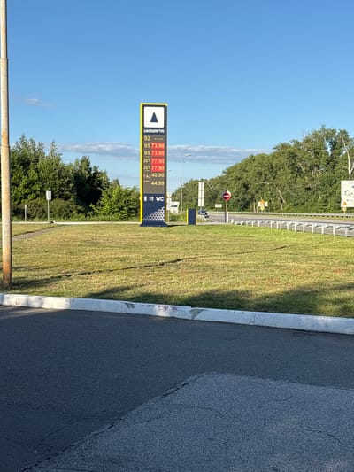 In the city of Nebelytsia, Ukraine, on a sunny day, a tall, illuminated price board for UKRNAFTA gas station stands prominently. The board displays fuel prices: 92 at "--.--", 95 at "73.90", another 95 at "73.90", ДП at "77.90", another ДП at "77.90", ГАЗ at "40.90", and AdBlue at "44.99". Below the fuel prices, icons for men's and women's restrooms and a "WC" sign are visible. The gas station is situated next to a grassy area with a paved road and a highway in the background. A metal pole, possibly a lamppost, stands to the left of the scene. The sky is clear blue with scattered white clouds. The overall environment appears to be a roadside service area.