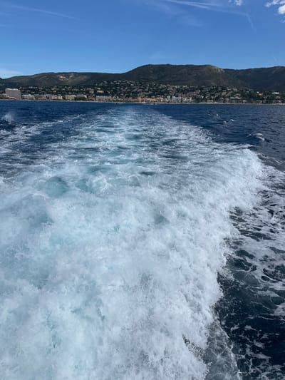 A wide-angle, low-angle, over-the-shoulder shot captures the wake of a boat moving away from the shore at Le Lavandou, France. The foreground is dominated by the churning, foamy white water of the wake, which trails off into the deep blue of the Mediterranean Sea. In the middle ground, a picturesque coastal town lines the base of a verdant, mountainous landscape. Buildings of various sizes and colors dot the hillside, interspersed with lush greenery. A clear blue sky with a few wispy clouds stretches across the horizon, indicating a bright, sunny day. The scene is devoid of people.