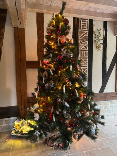 The image depicts an indoor Christmas decoration scene in Bailleul-la-Vallée, France. The environment features a room with light-colored plastered walls intersected by prominent dark wooden beams, indicative of a half-timbered architectural style. A section of the wall to the right displays a decorative pattern constructed from horizontal and diagonal brick-like elements between two vertical timbers. The floor consists of light-colored, square tiles.
Centrally, a fully decorated Christmas tree, estimated to be approximately 2.5 meters in height, stands illuminated. The tree is adorned with numerous small, multi-colored string lights, predominantly warm white and red. Its branches display a diverse collection of ornaments, including white candle-shaped lights clipped to the branches, red and white striped candy canes, various spherical baubles (some solid-colored, others patterned), miniature animal figurines (such as deer, squirrels, and birds), small house models, a rocking horse, and a mask.
At the base of the tree, to the left, a lighted miniature Christmas village display is present, featuring small buildings and figures illuminated by warm yellow lights. Under the front-right portion of the tree, a red and gold model train is positioned. An ornate, gold-colored two-candle wall sconce is mounted on the wall to the right of the tree. No individuals are present in the scene.