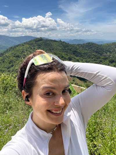 A woman is taking a selfie outdoors with a lush green mountain landscape and a blue sky with white clouds in the background. She is smiling and her right arm is raised behind her head. She is wearing a white zip-up jacket, sunglasses on top of her head, and black earbuds. The location is the city of Son Dien, Vietnam.