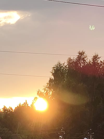 The image depicts a sunset over a natural landscape in Russkiye Dali, Russia. The sun, a bright orb of light, is positioned low in the sky, casting a warm glow over the scene. Silhouetted against the vibrant sky are the tops of numerous trees, their branches reaching upwards as if to embrace the fading light. The sky itself is a canvas of soft, pastel colors, transitioning from a pale yellow near the horizon to a muted grey higher up, with scattered clouds adding depth and texture.
In the upper right portion of the sky, a faint, blurry object with a greenish luminescence is visible. Its shape is indistinct, appearing somewhat circular with internal patterns, suggesting it could be an anomaly captured by the camera or a distant light source. The overall atmosphere is serene and tranquil, indicative of a peaceful end to the day. The presence of power lines stretching horizontally across the frame suggests a proximity to human habitation or infrastructure within the natural setting. The time of day is clearly sunset due to the low angle of the sun and the warm, diffused light. The weather appears clear, with only a few clouds in the sky. No people are visible in the image. There is no visible text.