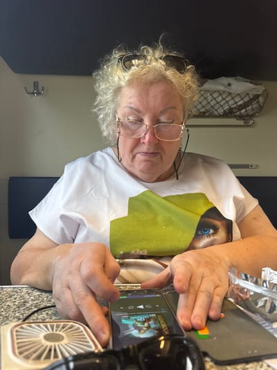 A medium shot captures a Caucasian woman with fair, curly hair, wearing glasses, and a white t-shirt with a large green graphic featuring a blue eye, seated at a table. Her hands are engaged with a smartphone, which rests on the table next to a small white fan and a silver foil packet. The background suggests an indoor, likely enclosed space, possibly a train compartment, with a dark ceiling and a wall featuring a coat hook. The overall lighting is dim. The image is taken from a slightly downward angle, focusing on the woman and her immediate surroundings. The woman's expression is neutral as she interacts with her phone. The setting appears to be within the city of Novye Biriuli, Russia.