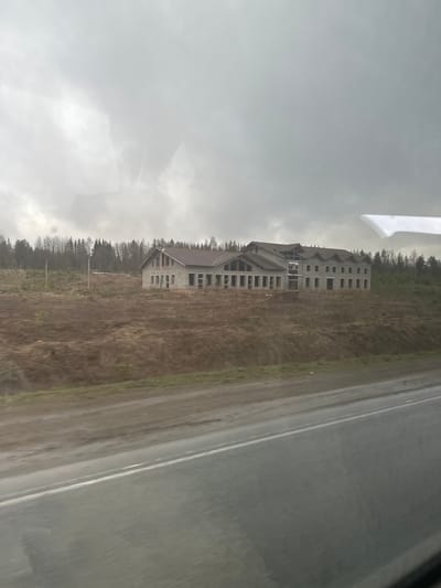 The image depicts an unfinished concrete building, likely a hotel or apartment complex, located in Bolgury, Russia. The structure is multi-storied and shows exposed concrete framing for windows and walls. The surrounding environment appears to be a rural area with a dirt field and a line of trees. An asphalt road runs alongside the building's property. The sky is overcast, suggesting a cloudy or rainy day. The image appears to be taken from inside a vehicle, possibly a bus or car, based on the reflections and the perspective.