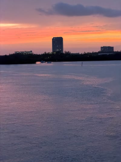This is a photograph of a sunset over a body of water in Bal Harbour Village, Florida, United States. The sky is a vibrant mix of orange, pink, and purple hues, with some darker clouds visible towards the top. In the distance, a silhouetted skyline features several buildings, including a tall, modern skyscraper and a lower-rise building. The water in the foreground is calm, reflecting the colors of the sky. A few boats are visible on the water near the shore. The overall impression is one of tranquility and natural beauty.