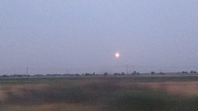The image depicts a hazy, twilight or dawn scene over a rural landscape, possibly in or around Łagów, Poland. A faint, glowing circular object, likely the moon or sun due to the atmospheric conditions, is visible in the upper center of the frame. The foreground consists of a field with a mix of muted greens and browns. In the middle ground, a line of trees and vegetation stretches across the horizon, interspersed with what appear to be utility poles and possibly power lines. The sky is a pale, uniform gray-blue, contributing to the overall soft and diffused lighting. No people or specific man-made structures are clearly identifiable, suggesting an open, natural environment.