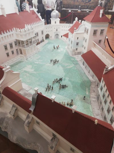 An elevated photograph captures a detailed architectural scale model of Mir Castle, situated in Мірскі сельскі Савет, Belarus, presented indoors within a museum environment. The model consists of multiple light-toned buildings featuring prominent red tile roofs, a central gateway with an arched entrance, and various towers, including one topped with a silver dome. The central courtyard, depicted with a textured, light-teal surface, hosts numerous miniature figures arranged in a historical battle formation. These figures comprise groups of infantry, cavalry, and artillery pieces (cannons), scattered across the courtyard simulating combat. Several partially visible adult spectators are positioned in the background, observing the exhibit. The surrounding environment includes dark wood display elements, a patterned tapestry on a wall, and red velvet rope barriers.