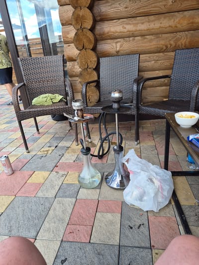 The image depicts a ground-level outdoor setting, likely a patio or veranda, adjacent to a wooden log cabin structure. The ground is paved with interlocking rectangular tiles in various shades of grey, pink, and yellow. In the foreground, two human legs are visible, suggesting a person is seated or lying down.

Several objects are arranged around the area. Two wicker chairs are placed on the tiled ground. On one chair, a crumpled green cloth is visible. In front of the chairs, two hookahs are set up. The hookahs are made of glass and metal, with black hoses. One hookah has a metal bowl on top. A silver can, possibly a beverage, is lying on the ground near the chairs.

A small, dark-colored metal table is positioned to the right of the hookahs. On this table, a white bowl containing what appears to be potato chips is placed. To the right of the table, a white plastic bag filled with items is situated on the tiled ground. A partial view of a person wearing a camouflage-patterned shirt and dark shorts is visible on the far left of the image, with their back to the camera. Reflections of the sky and a wooden structure are visible in the glass windows behind the chairs.

The context of Kosshy, Kazakhstan, is not directly deducible from the visual elements within the image itself. However, the presence of outdoor seating, hookahs, and a relaxed atmosphere suggests a recreational or hospitality setting. The wooden cabin architecture could be indicative of a dacha (summer house) or a similar type of recreational dwelling common in some regions. The sunny weather suggested by the bright lighting implies a daytime scene.