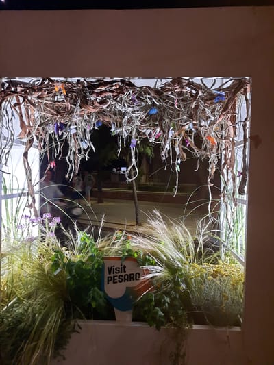 The image shows an outdoor art installation featuring dried branches and vines suspended from a white frame, adorned with numerous colorful, butterfly-shaped decorations. Beneath this overhead display, a lush arrangement of plants and grasses is visible, including tall, feathery ornamental grasses, bushy green foliage, and clusters of small yellow flowers. A white sign with the text "Visit PESARO" is placed among the plants. In the background, through the frame of the installation, several people are seen walking in what appears to be a public space at night, with some streetlights and trees visible. The overall scene suggests a festive or decorative display in the city of Pesaro, Italy, likely in a public area given the presence of people and street lighting.