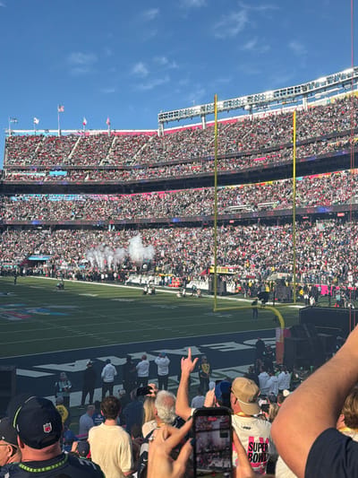A football stadium packed with spectators during a game, featuring goalposts and field markings. Smoke billows on the field. Foreground elements include onlookers, some holding phones to capture the event. One spectator wears a "SUPER BOWL" shirt. The scene captures the energy and excitement of a live sporting event.