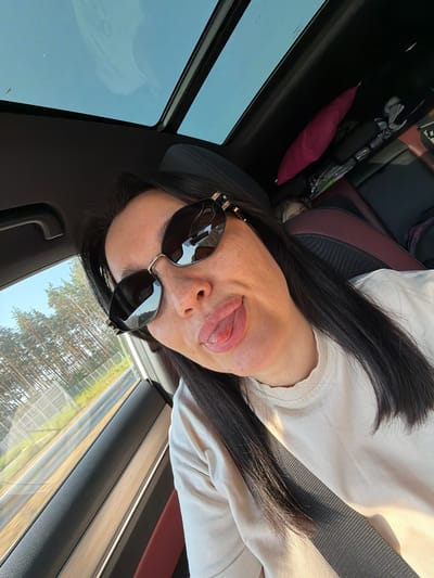 A woman with dark hair and sunglasses is sticking her tongue out while in the passenger seat of a car. She is wearing a light-colored sweatshirt and a grey seatbelt is visible across her chest. The car has a sunroof that is open, revealing a blue sky. Outside the window, a fence and trees are visible, suggesting a rural or suburban roadside. The interior of the car appears to be dark leather with red accents.