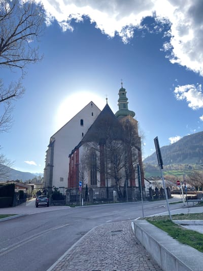 The image shows the church of St. Margaret in Sterzing-Vipiteno, Italy, from a low-angle perspective looking up. The church features a tall, green-roofed steeple and a white facade with red architectural details. The sun is positioned behind the building, causing a glare. A road runs alongside the church, with a few cars parked on it. A sidewalk lined with stone pavers is visible on the right. Bare tree branches are in the lower left corner and partially obscuring the church. The sky is blue with puffy white clouds. In the background, a hillside and buildings are visible. A traffic sign is on the side of the road.