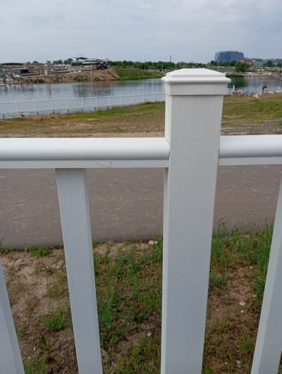 The image shows a white fence post in the foreground, with a white railing extending horizontally across the frame. Beyond the fence, there is a body of water, likely a river or lake, with a white walkway or bridge on its edge. In the background, there are buildings, trees, and a cloudy sky. The location is identified as Sol-Iletsk, Russia. The scene appears to be a public area, possibly a park or recreational area, with some scattered individuals visible in the distance, some appearing to be relaxing on lounge chairs. The overall impression is of a calm, outdoor setting.