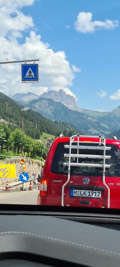 The image shows a rear view of a red Volkswagen Caddy LIFE with a bicycle rack mounted on the back. The license plate reads "MLK 1712". The car is stopped in traffic, indicated by the pedestrian crossing sign and traffic signals visible in the foreground. Behind the car, a majestic mountain range is visible under a bright blue sky with scattered clouds. The mountains are covered with lush green forests. The scene is set in a scenic location, likely a mountain road, in Vigo di Fassa - Vich, Italy, as suggested by the context of the license plate and the mountainous landscape.