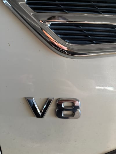 The image shows a close-up of the front of a vehicle, likely a car or SUV. The vehicle has a chrome grill with vertical bars visible on the left side of the frame. To the right of the grill, on the white exterior of the vehicle, are the letters "V8" in chrome. The "8" is positioned above the "V." The lighting appears to be bright and even, illuminating the chrome details and the white surface of the vehicle. There are no people visible in the image. The location, Hollywood Seminole Reservation, United States of America, is provided but not directly evident from the visual information in the photograph.