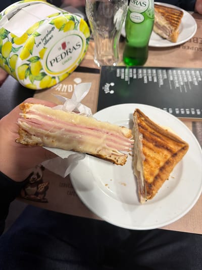 The scene depicts an indoor setting at Panda Café & Cervejaria, located at R. Manuel Luís Nogueira 229, 4420-192 Gondomar, Portugal. A human left hand holds a substantial, triangular portion of a grilled ham and cheese sandwich, revealing multiple layers of melted cheese and thinly sliced ham between toasted bread. Strands of melted cheese stretch from the held portion. A second, similar triangular piece of the grilled sandwich rests on a white ceramic plate to the right. The items are positioned on a brown placemat featuring a graphic of a panda holding a pizza slice, with partial text including "CERVEJA" and "CAFÉ." On the table, an empty clear drinking glass, a green bottle of Pedras Salgadas Natural mineral water, a lemon-themed cylindrical container branded "PEDRAS LIMÃO" (likely a napkin holder), and a black menu with white text are visible. The perspective is from slightly above, looking down at the table.