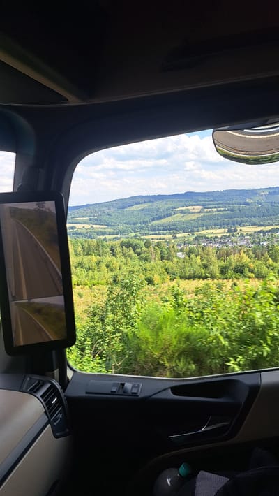 The image is taken from inside a truck cabin, looking out through the windshield and a side mirror. The exterior view shows a rolling, verdant landscape with a village nestled in a valley. Hills covered in dense green trees dominate the background, with some patches of cleared land and fields visible. The sky is partly cloudy with a bright, hazy sun. The truck cabin interior features dark gray and beige plastic trim, including an air vent, window controls, and a door handle. A side mirror, mounted on the exterior of the cabin, is visible on the left, reflecting a road. The image appears to be taken on a sunny day. The location is identified as being in the vicinity of Burbach, Germany.