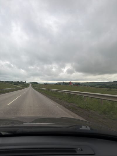 The image, taken from inside a vehicle, captures a view of an empty asphalt road stretching into the distance under an overcast sky. The road has a white line marking its center. On the right side of the road, there is a metal guardrail. Beyond the guardrail, a green grassy field extends to rolling hills in the background, dotted with trees. In the far distance, a cluster of buildings, possibly in a village or town, is visible, featuring some red structures. The weather appears to be cloudy, suggesting it could be late afternoon or an overcast day. The scene implies travel, with the road acting as the primary subject. No people are clearly visible, but a single red vehicle is seen on the road in the distance. The setting is rural, likely in Kendektamak, Russia, given the location context. The overall mood is tranquil, with the expansive road and cloudy sky creating a sense of solitude. There is no visible text in the image.