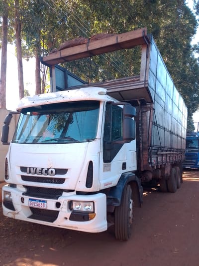 A white Iveco truck, license plate GVE5B68, is parked on a dirt road in São Gabriel do Oeste, Brazil, under a canopy of leafy trees. The truck has a large, canvas-covered cargo area and a visible load strapped to its roof. A blue truck, possibly a Volvo, is parked behind it. The scene is illuminated by sunlight filtering through the trees.