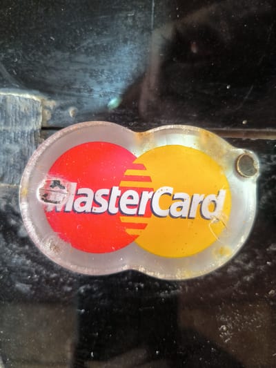 The image presents a close-up view of a heavily weathered and dirty transparent oval-shaped sticker or plaque displaying the MasterCard logo. The iconic logo features two interlocking circles: a vibrant red one on the left and a bright yellow-orange one on the right. The word "MasterCard" is printed in white letters with a thin blue outline across both circles, overlaid by yellow horizontal stripes on the red circle.
The sticker exhibits significant signs of age and wear. The transparent material is extensively scratched, scuffed, and covered in grime and debris, which dulls the logo's original colors. A prominent area of damage is visible over the 'M' in "MasterCard," partially obscuring the letter. A small, circular hole, likely for attachment, is present on the far right edge of the transparent plaque.
The logo is affixed to a dark, reflective surface that is equally dirty, covered in dust, smudges, and various marks. This background surface reflects light unevenly, with hints of lighter blue-grey and white patterns, suggesting a textured environment or strong reflections. A lighter, textured element, possibly a wall or structure, is faintly discernible behind the dark surface in the top and bottom left areas. The overall bright lighting suggests the image was taken during the daytime. The scene, likely located in Bateiiliek, Indonesia, captures a payment method sign showing considerable exposure and use.