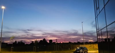 This media file depicts an outdoor scene at dusk, likely in Pabianice, Poland. The sky is a prominent feature, showcasing a gradient of colors from a clear, light blue at the top to vibrant pinks and purples near the horizon, indicating the sunset. Wispy clouds stretch across the sky, catching the last rays of sunlight.
In the foreground, a dark-colored car is parked on what appears to be a paved area or road. To the right of the car, a modern building with a glass facade reflects the colorful sky and surrounding environment. Several tall lampposts are visible throughout the scene, one in the foreground to the left, illuminated with a bright light, and another further back to the right.
Behind the car and building, a fence separates the paved area from a grassy field. Silhouetted trees and some smaller vegetation are scattered across the landscape. In the distance, power lines and structures are faintly visible against the twilight sky. A parking sign with a white 'P' on a blue background is positioned near the fence in the middle ground.
The overall mood is peaceful and serene, characteristic of a quiet evening. The illuminated lamppost suggests it is transitioning into night. No people are visible in the frame. The location cues like the parking sign and the architectural style of the building hint at an urban or suburban setting, possibly near a commercial or office area.