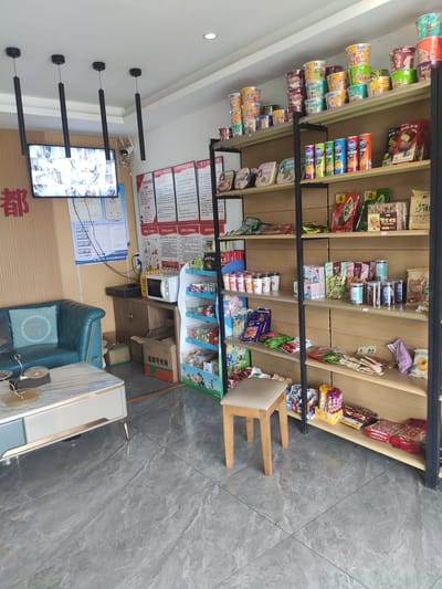 The image shows the interior of a small convenience store in Jianyang City, China. The store is well-lit and appears to be clean. In the foreground, a light-colored marble-patterned floor leads to a blue sofa with a patterned cushion. To the right of the sofa is a white and blue coffee table with a gold trim. Behind the sofa, a microwave sits on a dark wooden cabinet.

The main feature of the store is a tall shelving unit filled with various products. The shelves are stocked with an assortment of snacks, beverages, and other convenience items. On the upper shelves, there are stacks of cup noodles in colorful packaging. Below these, various canned drinks and packaged snacks are arranged. The shelves are made of light-colored wood, and the unit has a black metal frame.

On the wall to the left, a large screen displays surveillance camera feeds, indicating a security system is in place. Several signs with Chinese text are also visible on the wall, possibly related to store policies or regulations.

To the right of the shelving unit, a small wooden stool with a beige cushion is placed. The overall impression is of a functional and organized retail space.