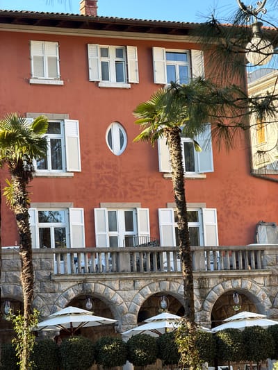The image depicts the facade of a building, likely a residential or commercial structure, in Grad Opatija, Croatia, based on its architectural style. The building has a red-toned exterior wall with multiple white-framed windows, some with shutters that can be opened or closed. An oval window is also present. A stone structure with arched openings and vegetation grows along the side of the building. Tall palm trees are visible in the foreground. The image appears to be taken in the afternoon sunlight.