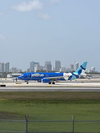 A JetBlue airplane is on the tarmac at an airport, with a city skyline in the background. The airplane is bright blue with the JetBlue logo on its side and a colorful design on its tail. It is a sunny day with a light blue sky and a few clouds. The airplane is positioned to the right of the frame, facing left, with its landing gear deployed. In the background, a dense cluster of modern high-rise buildings forms the city skyline. In the foreground, there is a grassy area and a chain-link fence. The image appears to have been taken in Dania Beach, Florida, given the distinctive skyline which is consistent with Fort Lauderdale's skyline, located in close proximity to Dania Beach.