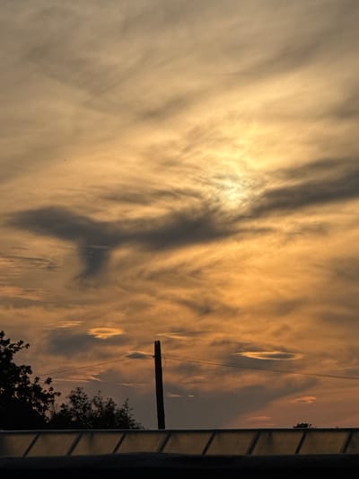 The image captures a sunset sky over Birgila, Russia, with the sun partially obscured by clouds, casting a warm orange and yellow glow. Wispy, dark clouds form an unusual shape resembling a figure or creature in the upper center of the frame. In the foreground, silhouetted against the sky are the tops of trees and a utility pole with wires. Below this, a dark structure with a translucent, angled roof runs horizontally across the bottom of the image. The overall scene depicts a dramatic and somewhat eerie natural phenomenon at dusk.