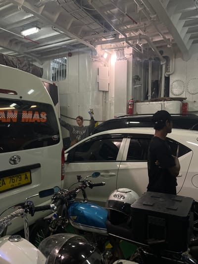 A photograph taken inside a vehicle ferry, likely in the city of Gunung Malang, Indonesia, depicts a crowded interior filled with vehicles and a few individuals. The ferry's interior is utilitarian, with exposed pipes and conduits overhead, and white painted walls. In the foreground, several motorcycles are parked, including a blue and white one with chrome accents. A white car with tinted windows is parked next to the motorcycles. Further back, a white van with orange lettering that reads "AN MAS" is visible. A woman in a grey sweatshirt is standing with her arms raised, holding a bottle and a snack. A man wearing a black t-shirt and a black baseball cap is standing to the right, facing away from the camera. The scene is dimly lit, with artificial lighting from overhead fixtures. The overall impression is one of a working ferry, transporting vehicles and passengers.