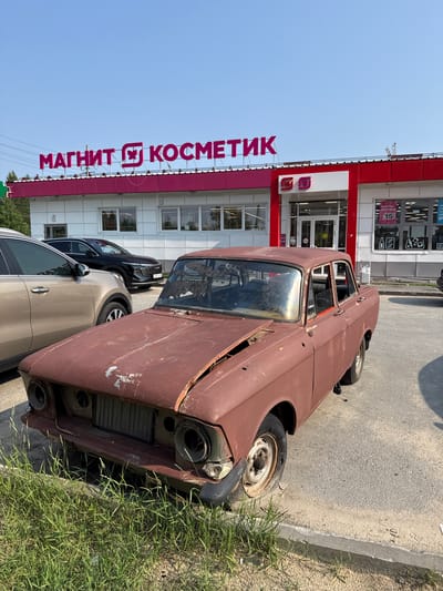 A rusty, abandoned car is parked on the side of a road in front of a Magnit Kosmetik store. The car is a dark reddish-brown and appears to be in poor condition, with visible rust and missing parts, including headlights and a front bumper. Its doors are open, and the interior is visible, though it is difficult to discern details.
In the background, the Magnit Kosmetik store has a bright red awning and a white building facade with several windows. Large pink lettering on the side of the building spells out "МАГНИТ КОСМЕТИК". Other cars are parked in front of the store.
The scene is set outdoors on a clear, sunny day. The presence of tall grass growing by the side of the road suggests an area that is not heavily maintained. The overall impression is one of urban decay or neglect, with the old car juxtaposed against the modern storefront. The time of day appears to be daytime, given the strong sunlight and shadows. There are no people visible in the image.