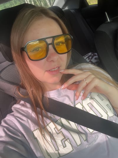A young Caucasian woman with long, light brown hair is shown from the chest up, sitting in the passenger seat of a car. She is wearing large, black-framed sunglasses with yellow lenses and a light purple t-shirt with a white and green graphic print. A black seatbelt is visible across her chest. Her right hand is raised to her face, with her index finger lightly touching her lips. She is wearing a thin gold ring on her left ring finger and has light pink, glittery nail polish on her fingernails. The background shows the interior of a car, including the headrests of the seats and a glimpse of the exterior through the windows, suggesting a daytime setting with trees and possibly a road. The image has a warm, slightly overexposed quality. The location is in the city of Brodokalmak, Russia.