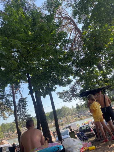 This is a horizontal, eye-level, outdoor photograph taken in the city of Khotyn, Ukraine. The scene depicts a sunny day at a lake or riverbank, with dense foliage from oak and pine trees framing the upper portion of the image. In the foreground, a shirtless man is seated, facing away from the camera. To the right, a young girl in a yellow outfit and two shirtless men are standing near a dark, angled structure. In the midground, a body of water is visible, with people swimming and several cars and tents lining the shore, suggesting a recreational area. The overall atmosphere is casual and summery.