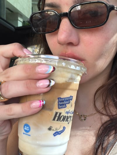 A close-up, slightly angled shot shows a person holding a clear plastic cup filled with a light brown, creamy beverage, likely an iced coffee. The person's face is partially visible, with one eye behind dark-rimmed sunglasses. Their lips are slightly parted, suggesting they are either about to drink or are in the process of drinking from the cup. The person has long, dark hair and is wearing a thin gold necklace with a circular pendant and a small diamond.
The cup has several stickers on it. One prominent sticker features the word "Honey" in large, black, stylized letters. Above it, in smaller text, are the words "cafe &". To the left of "Honey" is text that reads "Your everyday handmade break." To the right of "Honey" are two smaller stickers: one with a bee and the text "Bibi app" and another blue circular logo with "ec" inside, and "@earthecups" below it. The beverage appears to have ice and possibly whipped cream or a similar topping.
The person's nails are neatly manicured with a French tip design featuring colorful stripes of light blue, yellow, brown, and pink. They are wearing a gold ring on their left hand. The background is slightly out of focus but suggests an outdoor setting, possibly a car interior, given the visible window reflections and what appears to be a car's dashboard in the lower left. The lighting indicates it is daytime, with bright sunlight casting shadows. The overall mood seems relaxed and casual. The location is identified as South Miami, USA.
