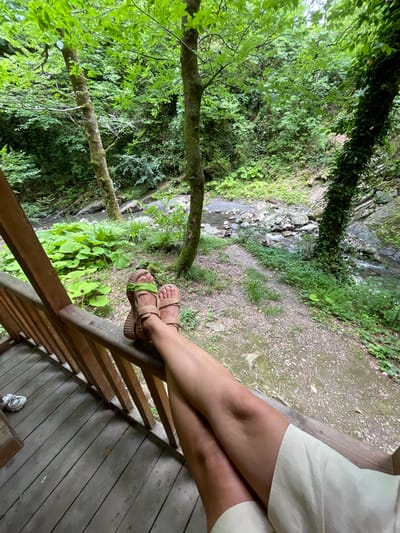A person's legs are crossed, resting on a wooden railing. They are wearing light-colored shorts and tan sandals with green accents. The scene is a lush, green forest with a stream flowing through it, visible in the background. The sun appears to be up, suggesting daytime. The environment is natural and serene, with a focus on the person's relaxed pose and the surrounding greenery.