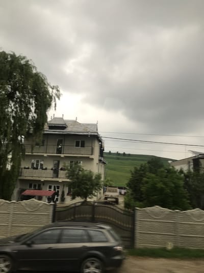 This image captures a street scene in Cristești, Romania, under an overcast sky. A multi-story, light-colored residential building with a complex, dark-tiled roof featuring multiple gables and decorative finials stands prominently in the mid-ground. The building has at least two visible floors with balconies, each equipped with dark metal railings. On the top balcony, a faint silhouette of a person is visible. Potted plants adorn the railing of the lower visible balcony. Below this balcony, a red awning or canopy extends outwards.
In the immediate foreground, a dark-colored station wagon, blurred by motion, travels from left to right. A decorative modular concrete fence, light grey with circular patterns and horizontal grooves, separates the road from the property. A dark, ornate metal gate, partially open, is situated to the right of the concrete fence.
To the left, a large tree with cascading foliage partially obscures the building and the sky. To the right of the main building, a lush green hillside rises in the background, topped by a sparse line of small trees. Utility lines stretch horizontally across the middle of the frame, passing in front of the house and the hillside. Parts of other residential structures are visible, including one to the far right with a solar panel on its roof and a white pickup truck parked nearby with laundry drying on a clothesline. The ground in the foreground appears to be an unpaved shoulder or dirt path.