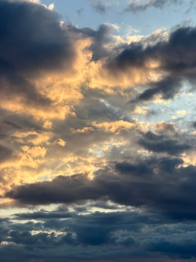 The image presents a sky at what appears to be either sunrise or sunset, given the light present. Dominating the frame are dense cloud formations. On the left and center of the image, clouds are illuminated with a golden hue, suggesting the light source is positioned behind them. In contrast, clouds on the right side appear darker, exhibiting shades of blue and gray. The sky visible between the clouds displays lighter tones, hinting at a source of illumination above the horizon. There is no specific information given that confirms the location is in the city of Brežine, Montenegro.