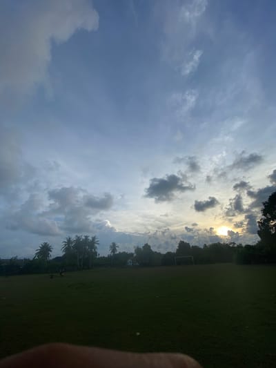 The image captures a partially rotated view of a sports field, likely a soccer field given the presence of a goalpost visible on the left side of the frame. The field is covered in lush green grass, with a few scattered pieces of litter visible on the ground. The background is a dense line of trees, with several prominent palm trees interspersed, suggesting a tropical or subtropical location. Some buildings are partially obscured by the foliage.
The sky is a prominent feature, with dramatic cloud formations. The sun is partially visible, breaking through the clouds and casting a bright glow, indicating either sunrise or sunset. The lighting suggests it is either early morning or late afternoon. The weather appears to be changing, with a mix of bright sunlight and dark, heavy clouds.
The presence of a few figures on the field suggests some activity, possibly a game or practice, though they are small and indistinct due to the distance and lighting. A human finger is partially visible in the bottom right corner, indicating the perspective of someone taking the photograph.
The overall mood is one of natural beauty, with the dynamic sky creating a sense of drama and atmosphere. The location context of Keudeublang, Indonesia, further supports the presence of palm trees and tropical vegetation. There is no visible text in the image.
