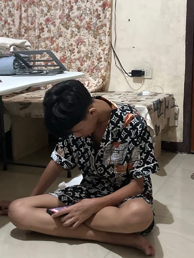A person with black hair is sitting cross-legged on the floor, looking down at a smartphone in their hands. They are wearing a black and white patterned pajama set with a repeating Louis Vuitton-inspired logo. The person is in a room with floral curtains and a table covered with a patterned cloth in the background. An electrical outlet with a charger plugged in is visible on the wall. The scene appears to be indoors, likely a bedroom or living area. The location is identified as Koronadal City, Philippines.