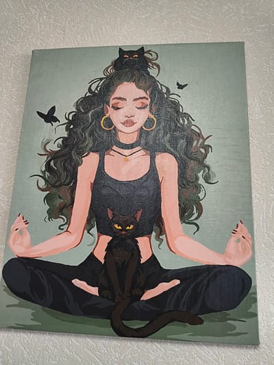 A painting depicts a woman meditating with two black cats and two black birds against a muted green background. The woman has long, dark, wavy hair and is wearing a black crop top and black pants. She is sitting in a lotus position with her eyes closed and hands resting on her knees. A black cat sits on her lap, and another black cat sits on top of her head. The birds are in flight around her. The painting is mounted on a canvas and displayed on a textured white wall. The location of Podkletnoe, Russia, is not depicted in the image.