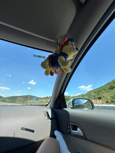 The image shows the interior of a car on a sunny day, with a plush toy dog hanging from the rearview mirror. The toy dog appears to be Chase from Paw Patrol, wearing a blue uniform and cap. The car is driving on a highway, with a view of green hills and a clear blue sky with some white clouds in the background. A road sign visible in the distance indicates "Kato Nevrokopi, Greece." The interior of the car shows the dashboard, door panel, and a person's leg and foot. The lighting suggests it is daytime.