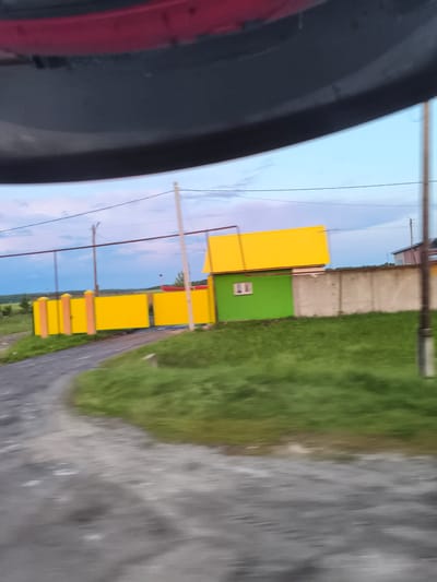 The image captures a blurry, outdoor scene during what appears to be daylight. In the foreground, a gray, uneven surface, likely a road or path, is visible, with patches of white or light-colored debris. Adjacent to this surface is a strip of vibrant green grass.

Dominating the mid-ground is a bright yellow fence, composed of multiple sections and punctuated by brick-colored posts topped with yellow caps. Behind the fence, a small, green building with a prominent yellow roof is situated. The building has a single, white-framed window. Further behind this structure, a light gray, concrete wall stretches horizontally.

In the background, the sky transitions from a light blue near the horizon to a slightly darker hue overhead, with hints of clouds. A power line runs horizontally across the sky, supported by tall poles. The overall impression is of a rural or semi-rural setting, possibly a residential area or property boundary. The location is identified as Трофимовка, Russia.