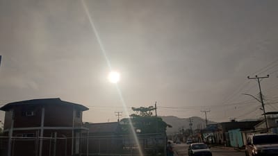 The image shows a street in Barcelona, Venezuela, under a bright, hazy sky with the sun high overhead. In the foreground, a two-story building with a dark roof and red brick or siding is visible, partially obscured by a chain-link fence. The street stretches into the distance, lined with low-rise buildings of varying colors and conditions. Power lines crisscross above the street. Several cars are visible on the road, with two cars in the foreground closest to the viewer. A person stands on the side of the road. In the background, a hill or small mountain is visible.