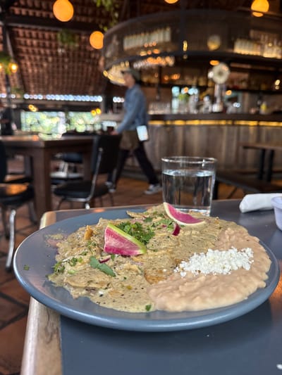The image shows a plate of food, likely chilaquiles, in a restaurant setting. The plate is light blue and contains two distinct sections: one with refried beans topped with crumbled white cheese and the other with tortilla chips covered in green sauce, garnished with cilantro and thin radish slices. A glass of water sits beside the plate.
In the blurred background, a bar area and dining space are visible. There is a person walking in the distance towards the middle of the image. The scene suggests a casual dining experience at a restaurant, potentially in La Tijera, Mexico, based on the request. The time of day appears to be either daytime or early evening, given the natural light.