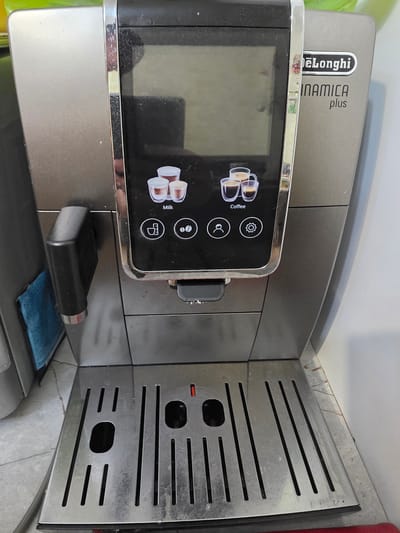 This image shows a close-up, angled view of a DeLonghi Dinamica Plus coffee machine, oriented vertically within what appears to be a domestic setting, possibly a kitchen. The machine is predominantly silver and black, with a prominent touchscreen display on the left side. The screen shows various icons, including three cups of coffee, a milk carton, a gear symbol, a bean symbol, and a measuring cup symbol. The DeLonghi and Dinamica Plus branding is visible on the machine's upper section.
To the right of the touchscreen is a stainless steel drip tray with horizontal slots. There is a small black component on the machine, possibly a milk frother attachment, resting on a blue cloth. The background is out of focus but suggests a light-colored wall and countertop. Some reddish-brown stains are visible on the stainless steel drip tray. The lighting appears to be artificial, suggesting an indoor environment. There are no people visible in the image. The overall impression is of a functional appliance in a typical home environment.
