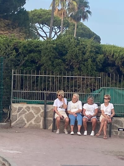 Four women are sitting on a bench outdoors in Playa Del Sol-Villacana, Spain. The woman on the far left is wearing a white t-shirt and light-colored shorts, with blonde hair. The second woman from the left has blonde hair and is wearing a white t-shirt and blue jeans. The third woman from the left has short grey hair and is wearing a white t-shirt and light-colored shorts. The woman on the far right has blonde hair and is wearing a white t-shirt and light-colored skirt. They are all looking in different directions. Behind them is a metal fence and a stone wall. In the background, there are trees and palm trees, suggesting a sunny, warm climate. The ground in front of the bench is a reddish-brown paved area.