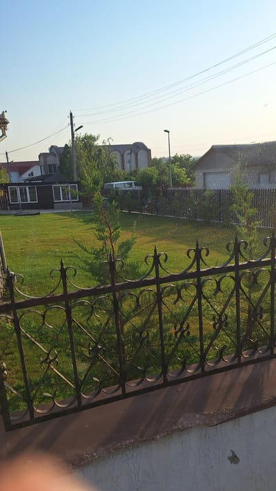 The image shows a residential property in Sărata-Galbenă, Moldova, on a clear, sunny day. In the foreground, a black wrought-iron fence runs horizontally across the bottom of the frame, partially obscuring a lush green lawn. Two small evergreen trees are visible behind the fence, growing on the lawn.
Beyond the lawn, a dark-colored building with large windows is situated on the left side of the property, likely a house or a garage. Further back, a white van is parked. A tall fence, also dark in color, runs along the back of the property, separating it from neighboring areas.
In the background, several houses with distinctive arched rooflines and brickwork are visible, suggesting a typical village or suburban architectural style in the region. Electrical wires and utility poles are strung across the sky, indicating the presence of infrastructure. A streetlamp stands near the center of the background. The sky is clear blue, with a slight haze near the horizon, and the sun is casting shadows, indicating it is likely morning or late afternoon. The overall scene depicts a tranquil and orderly residential setting.