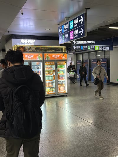 This image depicts an indoor public concourse within a metro station, identified as being in Yuhuatai District, Nanjing, China, by the presence of Nanjing Metro Line S1 and S3 signage and "Nanjing Specialty" branding. The environment features a polished, light-grey speckled floor and a white ceiling with recessed lighting.
In the left foreground, a male individual is observed from behind, wearing a black puffer jacket and olive-green trousers, with a black "sportporter" backpack. White wireless earbuds are visible in his ears. He stands facing two brightly lit, glass-fronted vending machines branded "南瓜先生" (Mr. Pumpkin), which display various bottled beverages and packaged items, including "南京特产" (Nanjing Specialty). An illuminated sign above the machines indicates "开门自取 / 扫码付款" (Open door and take / Scan code to pay).
In the mid-ground and background to the right, several individuals are present. One male subject walks from right to left, wearing a light-colored jacket, brown trousers, glasses, a light-blue face mask, and carrying a black shoulder bag. Other subjects are walking or standing further in the background. Overhead, a large blue and white directional sign points left to "1号线 Line 1" and "S1线 Line S1," and right to "3号线 Line 3" and "S3线 Line S3." Below this, another blue sign directs to "铁路出发(站)/售票/到达" (Railway Departure (Station)/Ticket/Arrival) and "乘车 1号线 3号线 出 2~6" (To Take Line 1 Line 3 Exit 2~6), indicating a connection to a railway station. The scene is evenly illuminated, casting faint reflections on the floor.
