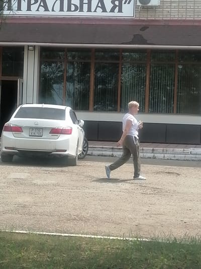 A blonde woman with short hair, wearing a white t-shirt and gray sweatpants, walks from left to right across a gravel lot. She is holding a small item in her right hand. Behind her, a white Honda Accord sedan is parked. The car has a license plate that reads "P 310 XK 125". In the background, a building with large windows and a sign with Russian text is visible. The sign appears to say "ТРАЛЬНАЯ". An air conditioning unit is mounted on the brick wall above the building's entrance. The scene appears to be in a commercial or public area. The ground is covered in gravel and there is a strip of green grass in the foreground. The weather is clear and sunny.