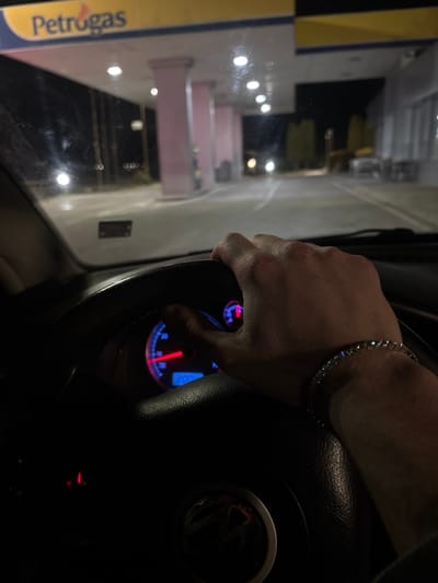 The image presents a nighttime view from the interior of a vehicle, parked or stopped at a Petrogas fuel station in Strumyani, Bulgaria. The driver's right hand, wearing a silver-toned bracelet with multiple clear, reflective stones, is positioned on the dark steering wheel, which bears a partially visible emblem resembling Volkswagen. Behind the steering wheel, an illuminated instrument cluster displays a speedometer with a red needle at a low reading and a blue digital display. Through the windshield, the brightly lit Petrogas canopy, distinguished by its yellow and blue signage, extends overhead. Below the canopy, several light pink support columns are visible, along with fuel pump islands and reflective ground surfaces. The area is illuminated by numerous circular white lights embedded in the canopy. In the midground to the right, a white building with an outdoor seating area is present. Distant blurred vehicle headlights and other ambient lights are visible against the dark background.